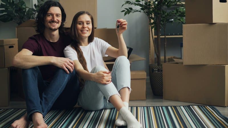 Casal sorridente sentado no chão do apartamento novo, entre caixas de mudança
